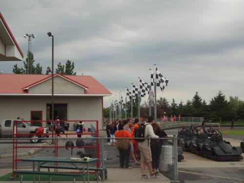 A go-kart track with people gathered near the entrance, checkered flags flying in the background under a cloudy sky.