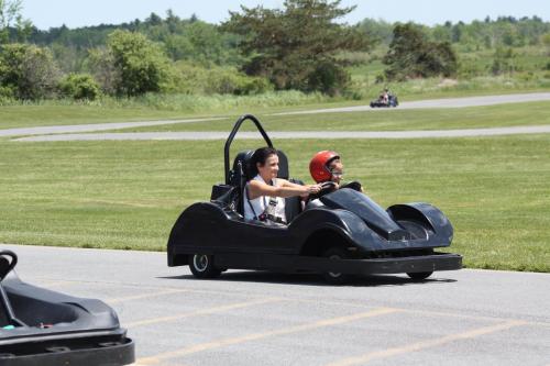 Two children race go-karts on a track surrounded by green grass and trees on a sunny day.