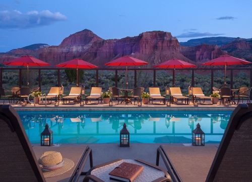 A serene pool area with lounge chairs and red umbrellas, set against a backdrop of mountains at dusk.