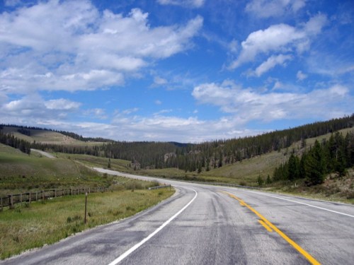 A winding road through green hills under a blue sky with scattered clouds. Trees line the roadside in the distance.