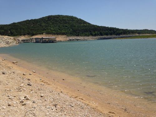 A serene lakeside view with a sandy shore, clear water, and a green hill in the background under a blue sky.