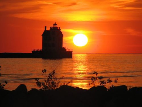 Silhouette of a lighthouse against a vibrant sunset over calm waters, with a glowing sun on the horizon.