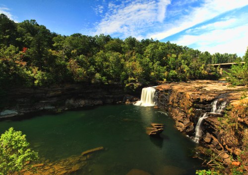 A serene waterfall cascades into a tranquil pool, surrounded by lush greenery and a clear blue sky.