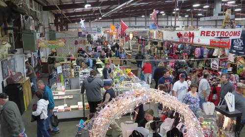 A bustling indoor market filled with shoppers browsing various stalls and displays, with festive decorations overhead.