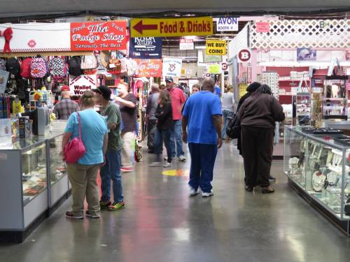 A busy indoor market with shoppers browsing various stalls and displays, featuring food and drink signs overhead.
