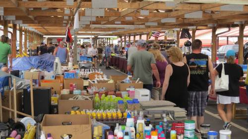 A bustling market with people browsing various stalls filled with goods and boxes under a wooden structure.