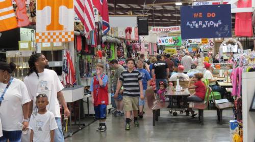 A bustling indoor market with shoppers, colorful flags, and various stalls selling goods and food.