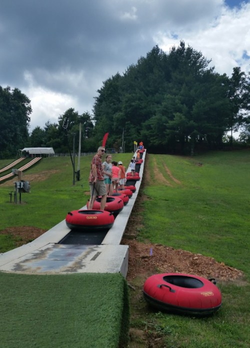 A group of people lined up on a grassy slope, preparing to ride inflatable tubes down a slide.