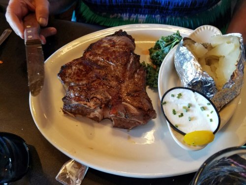 A plate with a large steak, a baked potato wrapped in foil, and a side of greens, with a knife resting nearby.