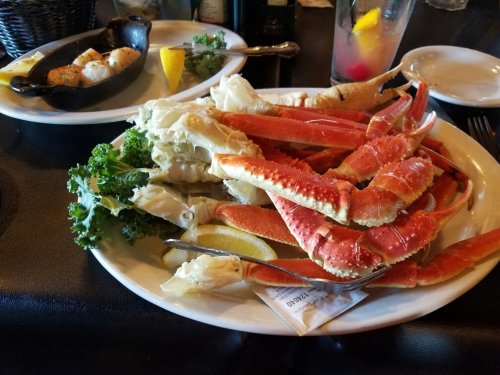 A plate of crab legs with lemon and kale, accompanied by a side of baked seafood and drinks.