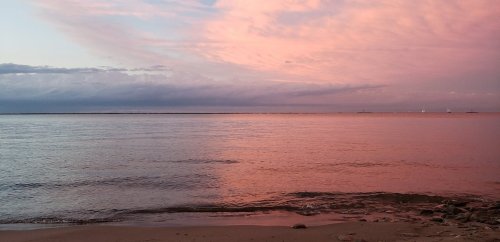 A serene beach at sunset, with soft pink and purple hues reflecting on calm water and a rocky shore.