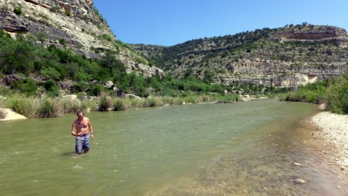 A man wades in a river surrounded by lush greenery and rocky hills under a clear blue sky.