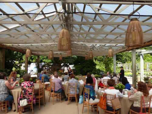 A group of people seated at tables under a canopy, enjoying a gathering in a garden setting.