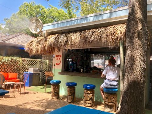 A tropical outdoor bar with thatched roof, stools, and a fan, featuring a person sitting at the counter.