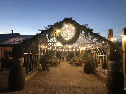 A festive greenhouse adorned with lights and greenery, surrounded by potted plants, at dusk.