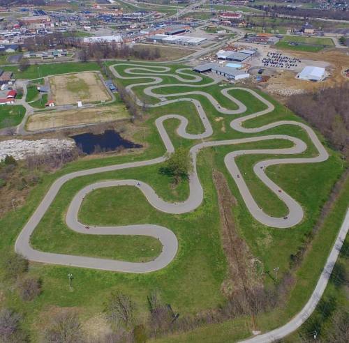 Aerial view of a winding go-kart track surrounded by green fields and a small pond, with a town in the background.