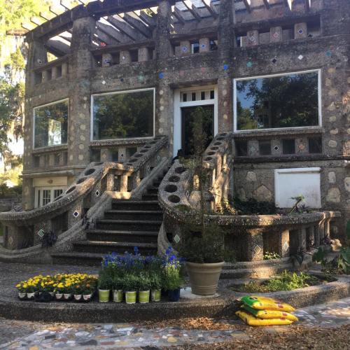 A unique, textured building with large windows, surrounded by colorful flowers and plants in pots.