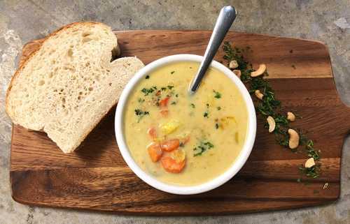 A bowl of creamy soup with vegetables, served with a slice of bread on a wooden board, garnished with herbs and nuts.