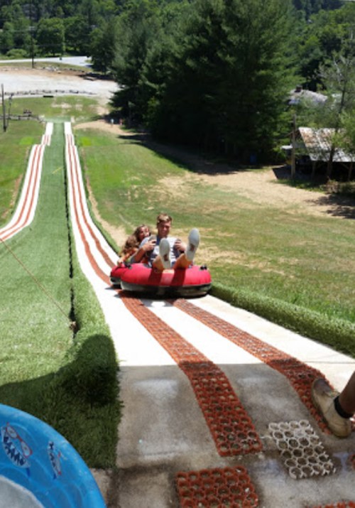 Two people sit in a red inner tube, sliding down a grassy slope at a tubing park on a sunny day.