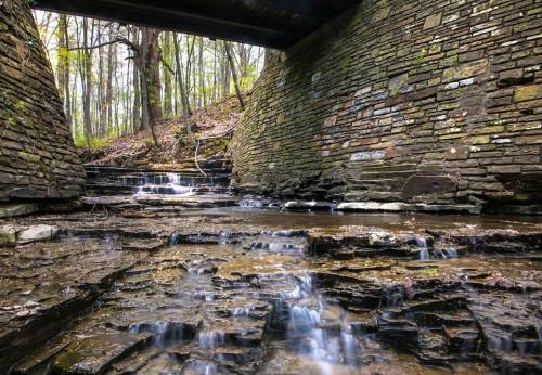 A serene stream flows over rocky terrain beneath a stone bridge, surrounded by lush green trees in a tranquil forest setting.