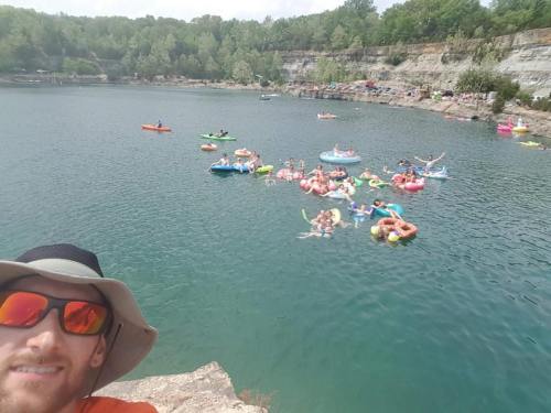 A man in a hat and sunglasses takes a selfie by a lake filled with people on colorful floats and kayaks.