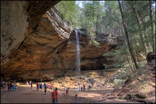 A scenic view of a waterfall cascading from a rocky overhang, with people exploring the area below in a lush forest.