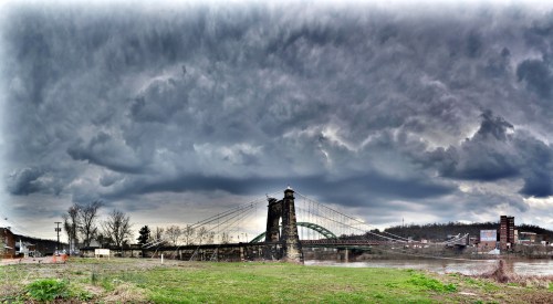A dramatic sky looms over a historic bridge, with lush grass in the foreground and industrial buildings in the background.
