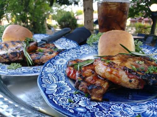 Grilled meat served on blue floral plates with rolls and a glass of iced drink, set outdoors with greenery.