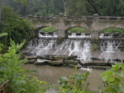 A stone bridge with arches over a cascading waterfall, surrounded by lush greenery and a calm river below.