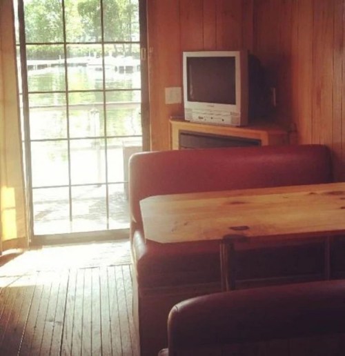 Cozy interior of a cabin with a wooden table, red seating, and a view of a lake through large glass doors.