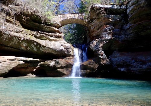 A serene waterfall cascades into a clear pool beneath a stone arch bridge, surrounded by lush greenery and rocky cliffs.