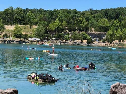 A sunny lake scene with people swimming, paddleboarding, and relaxing on floats among green trees and clear water.