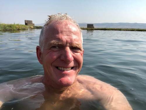 A smiling man with short gray hair is floating in calm water, surrounded by greenery and distant structures.