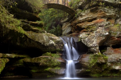 A serene waterfall cascades over rocks, surrounded by lush greenery and a natural stone arch above.