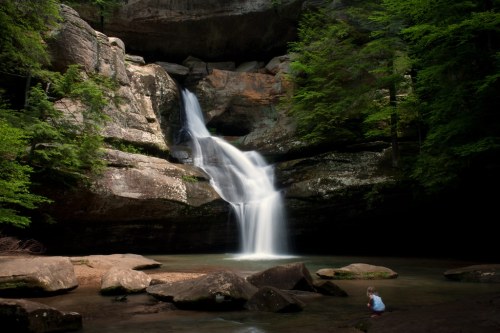 A serene waterfall cascades over rocks into a calm pool, surrounded by lush green trees and a child sitting nearby.