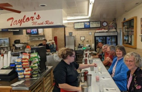 A diner scene at Taylor's Maid-Rite with a staff member serving drinks to a group of smiling customers at the counter.