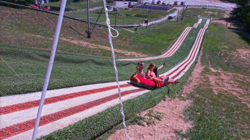 Two girls sit on a tubing slide, preparing to go down a grassy slope with a scenic view in the background.