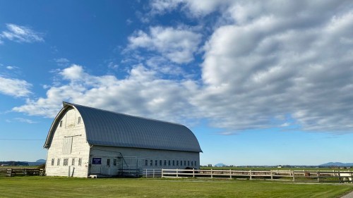 A large white barn with a metal roof stands in a grassy field under a blue sky with scattered clouds.