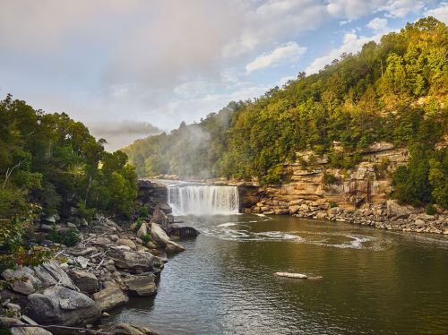 A serene waterfall cascades into a calm river, surrounded by lush green trees and rocky shores under a cloudy sky.