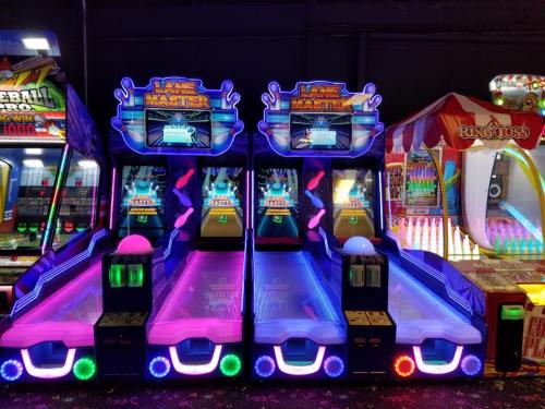 Brightly lit arcade game machines with neon colors, featuring skee-ball style gameplay and a carnival booth in the background.