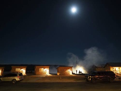 Night scene with cabins under a bright moon, smoke rising in the background, and silhouettes of people.