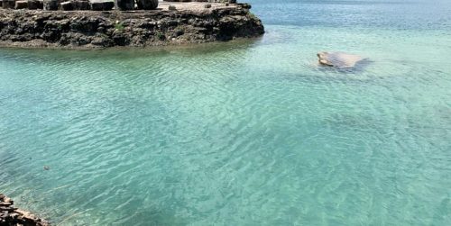 Clear turquoise water gently ripples around rocky shorelines under a bright blue sky.