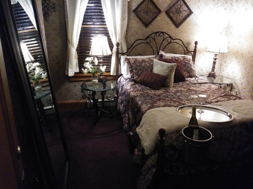 Cozy bedroom with a patterned bedspread, two lamps, a mirror, and a tray with champagne on a side table.