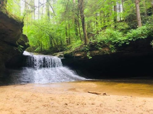 A serene waterfall cascades into a sandy area, surrounded by lush green trees and rocky cliffs.