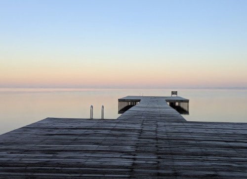 A serene wooden dock extends into calm waters, with a soft pastel sky at dawn or dusk.
