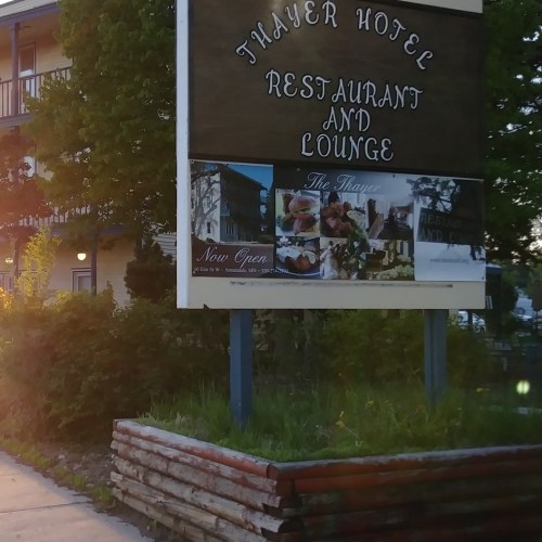 Sign for Thayer Hotel Restaurant and Lounge, with greenery and a building in the background, illuminated by sunlight.