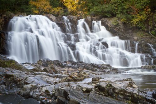 A serene waterfall cascades over rocky terrain, surrounded by autumn foliage and a calm river below.