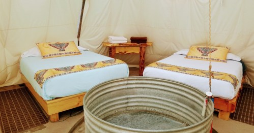 Cozy tent interior with two beds, decorative pillows, a wooden table, and a round metal basin in the center.