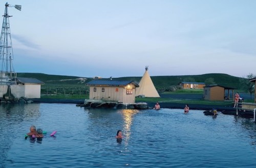 People swimming in a natural pool near wooden structures and a teepee, with a windmill in the background at dusk.