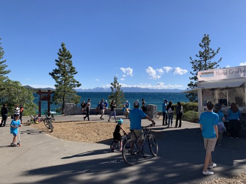 A busy lakeside area with people, trees, and a food stand, overlooking a blue lake and mountains in the background.